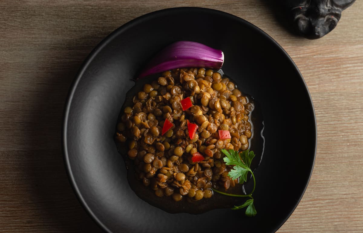 Cooked brown lentils with diced tomato, a wedge of red onion and a parsley sprig in a shallow black bowl, seen from above.