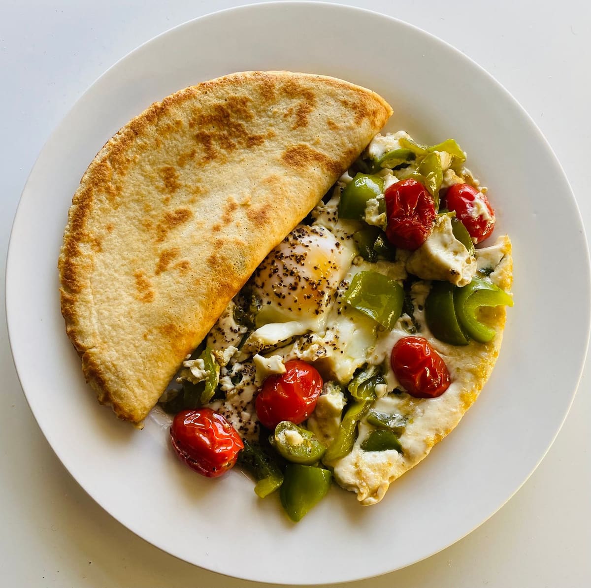 Eggs cooked with strips of green bell pepper, roasted cherry tomatoes, and crumbled white cheese on a white plate, with a folded flour flatbread beside them.