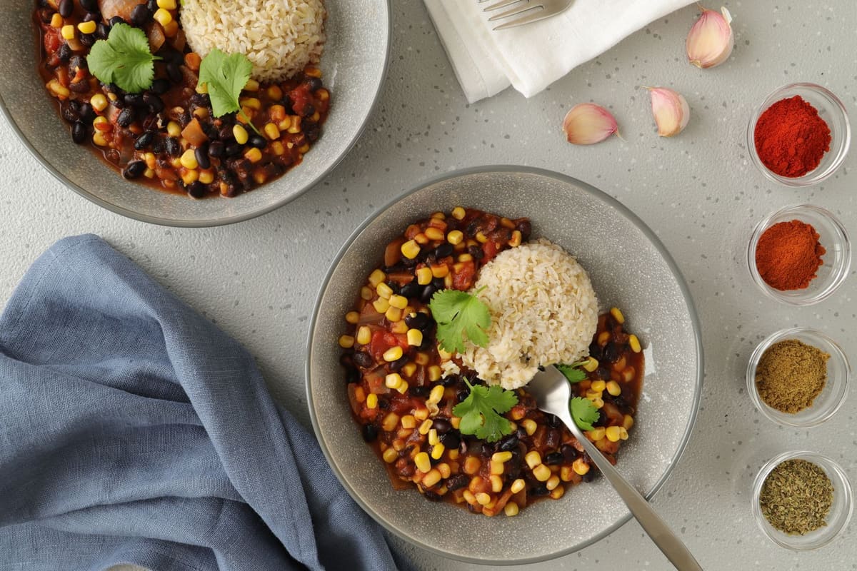 Two grey bowls of black bean and corn chili in a tomato red sauce, garnished with cilantro, with small dishes of ground spices beside them.