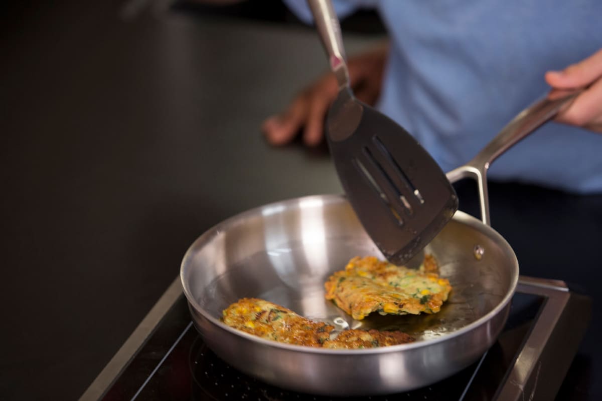 Golden corn and zucchini pancakes cooking in a stainless steel skillet, one lifted on a spatula to turn it.