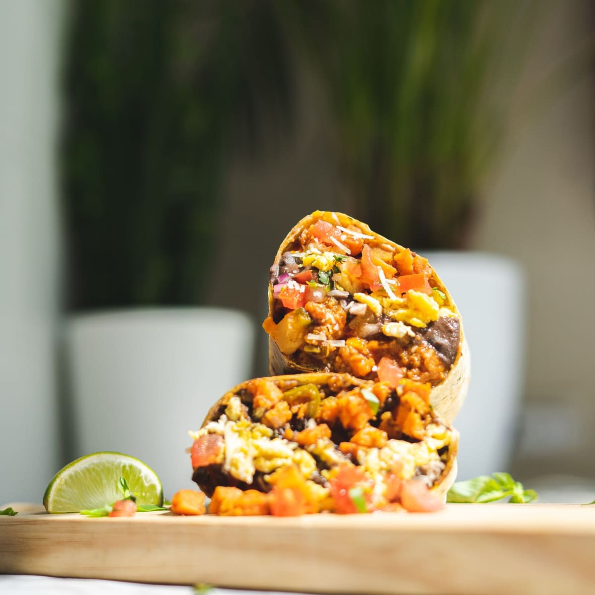 A flour tortilla burrito cut in half and stacked, the cut ends showing scrambled egg, black beans, diced potato, and tomato, with a lime wedge on the board.