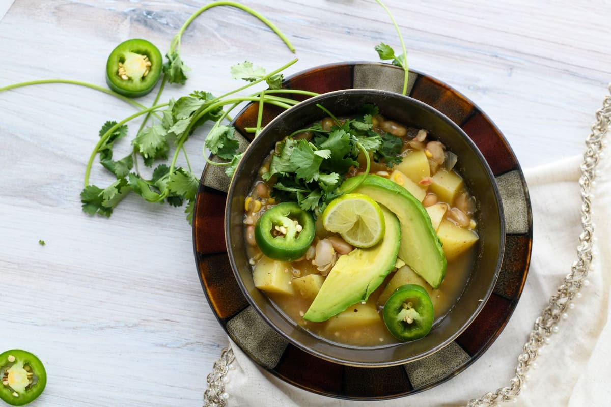 A dark bowl of brothy soup with chunks of potato and pale beans, topped with avocado slices, sliced jalapeno, and cilantro.