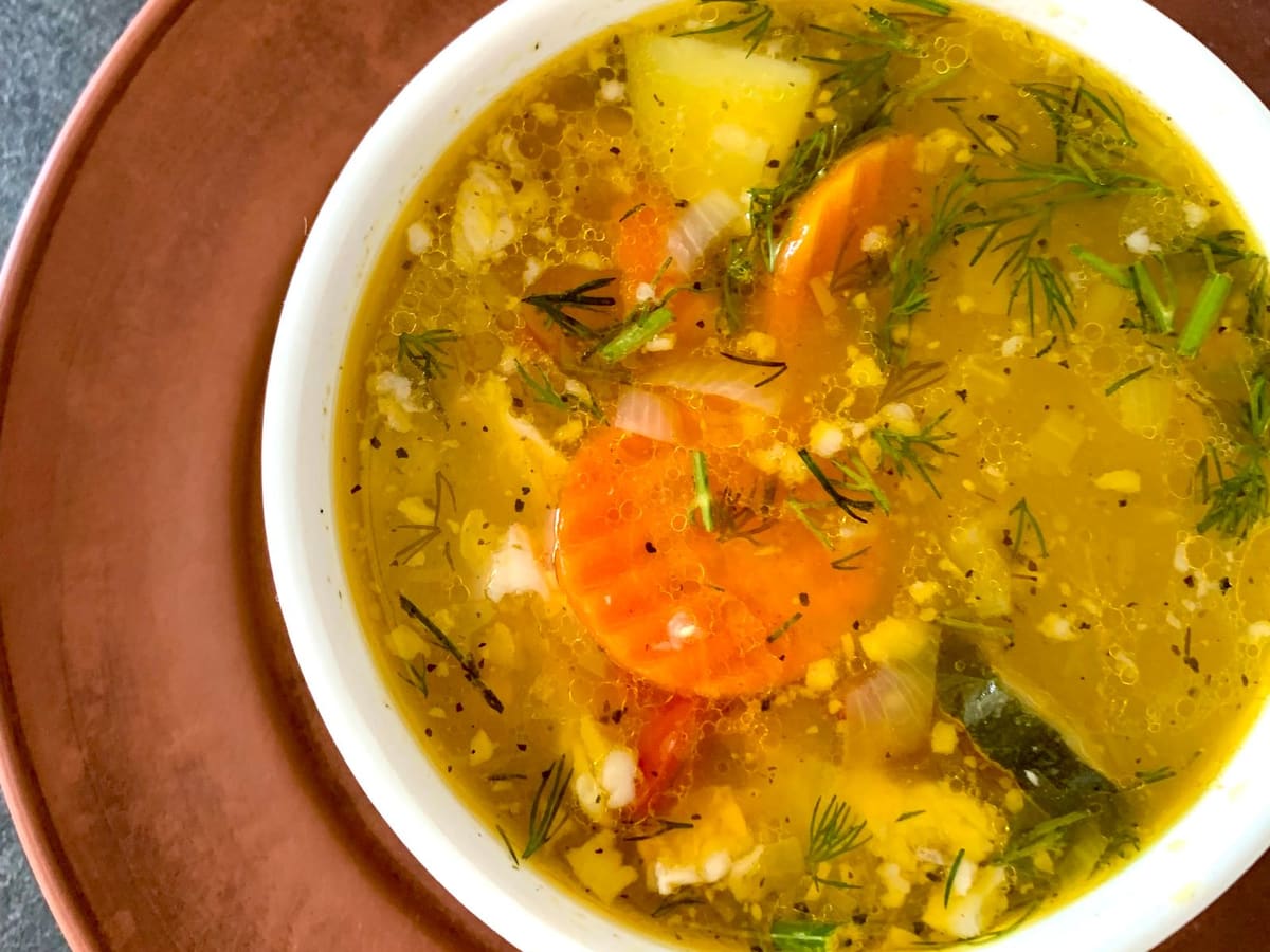 A white bowl of clear vegetable soup with chunks of potato, carrot slices, onion, and fresh dill, set on a terracotta plate.