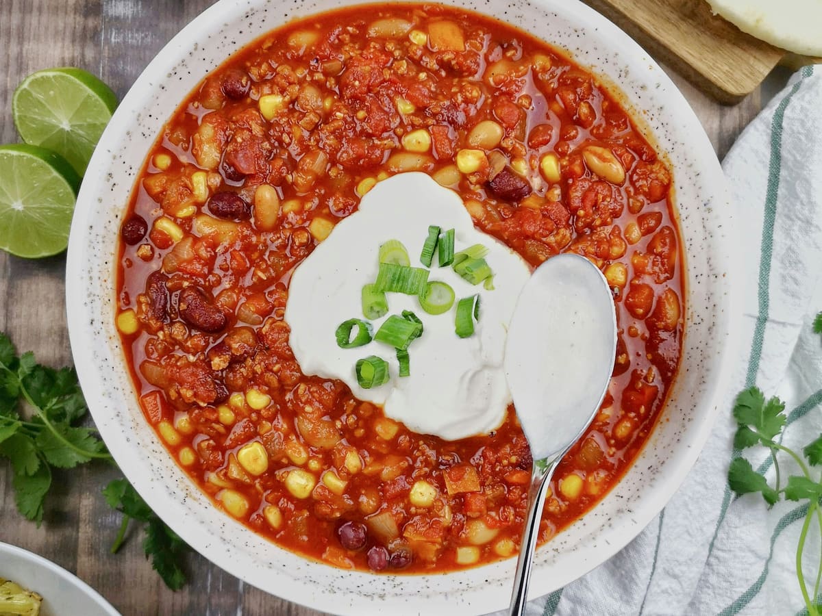 A bowl of bean chili with corn and tomato, topped with sour cream and sliced green onion, with a spoon resting in it and lime wedges and cilantro beside the bowl.