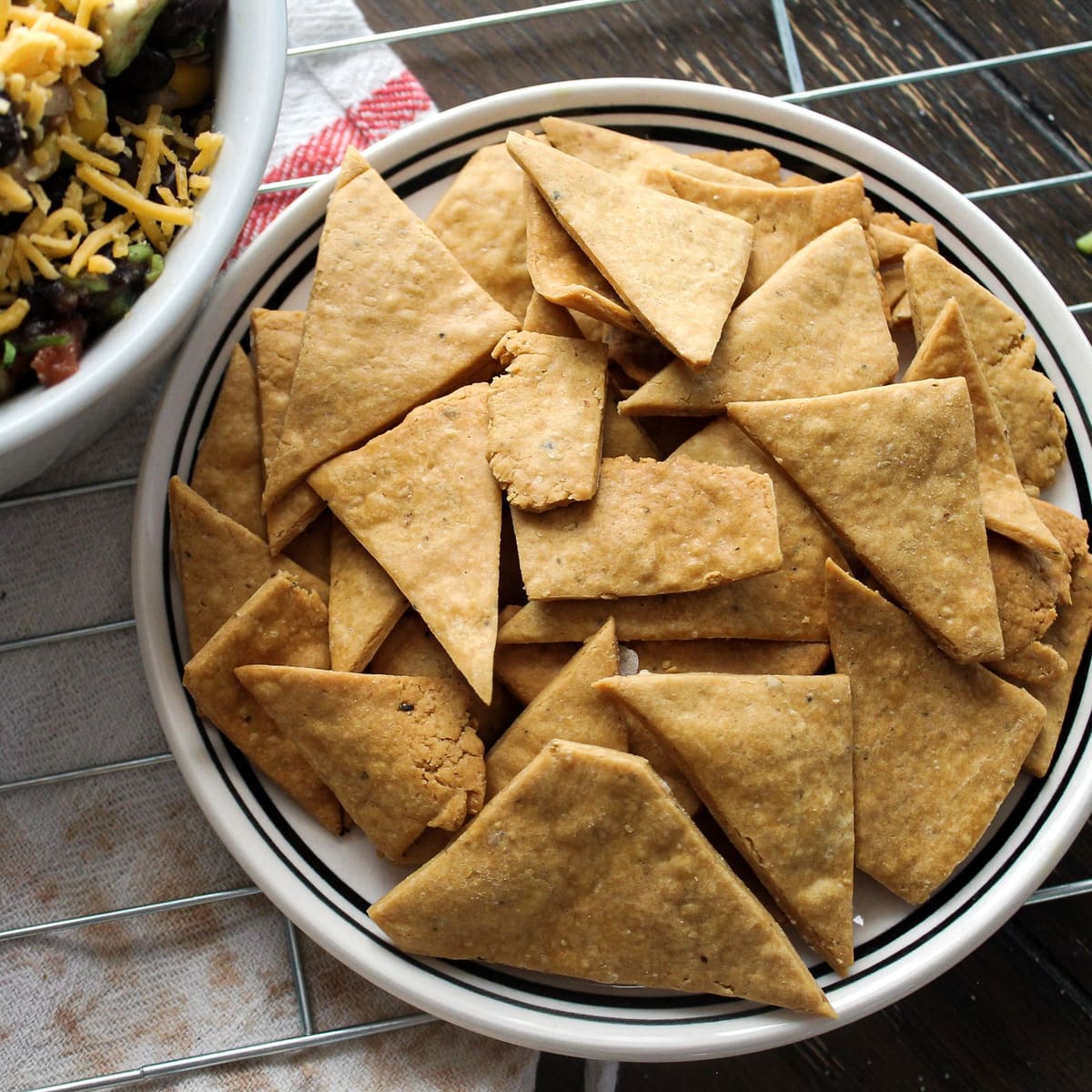 Baked tortilla triangles piled on a rimmed white plate on a wooden table, with a bowl of black bean and corn salsa at the edge.
