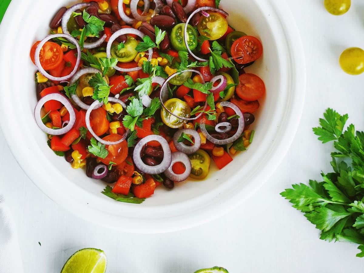 A white bowl of bean and corn salad, with dark beans, corn kernels, diced red pepper, halved cherry tomatoes, rings of red onion, and flat leaf parsley, and lime wedges on the table beside it.