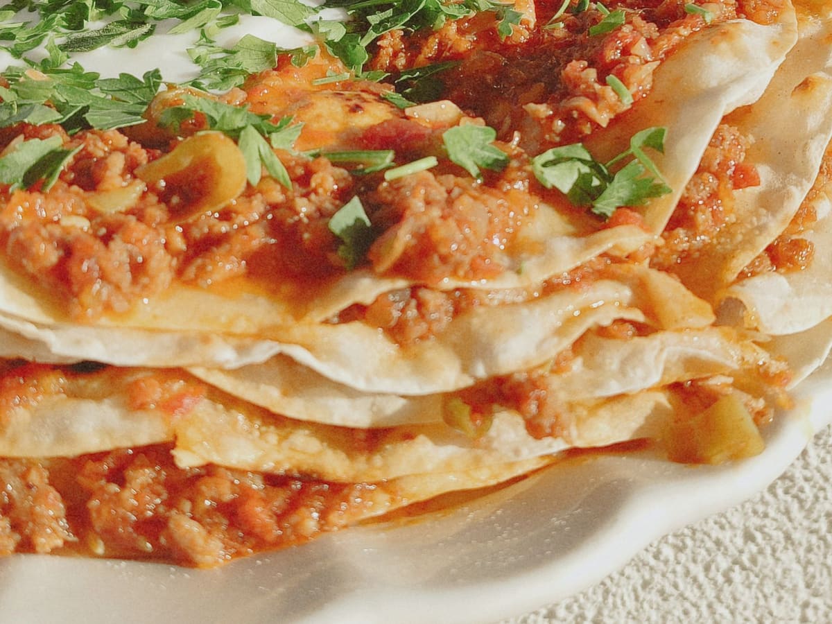 A close view of a baked dish of stacked tortillas layered with a tomato, pepper, and meat filling, scattered with chopped parsley.