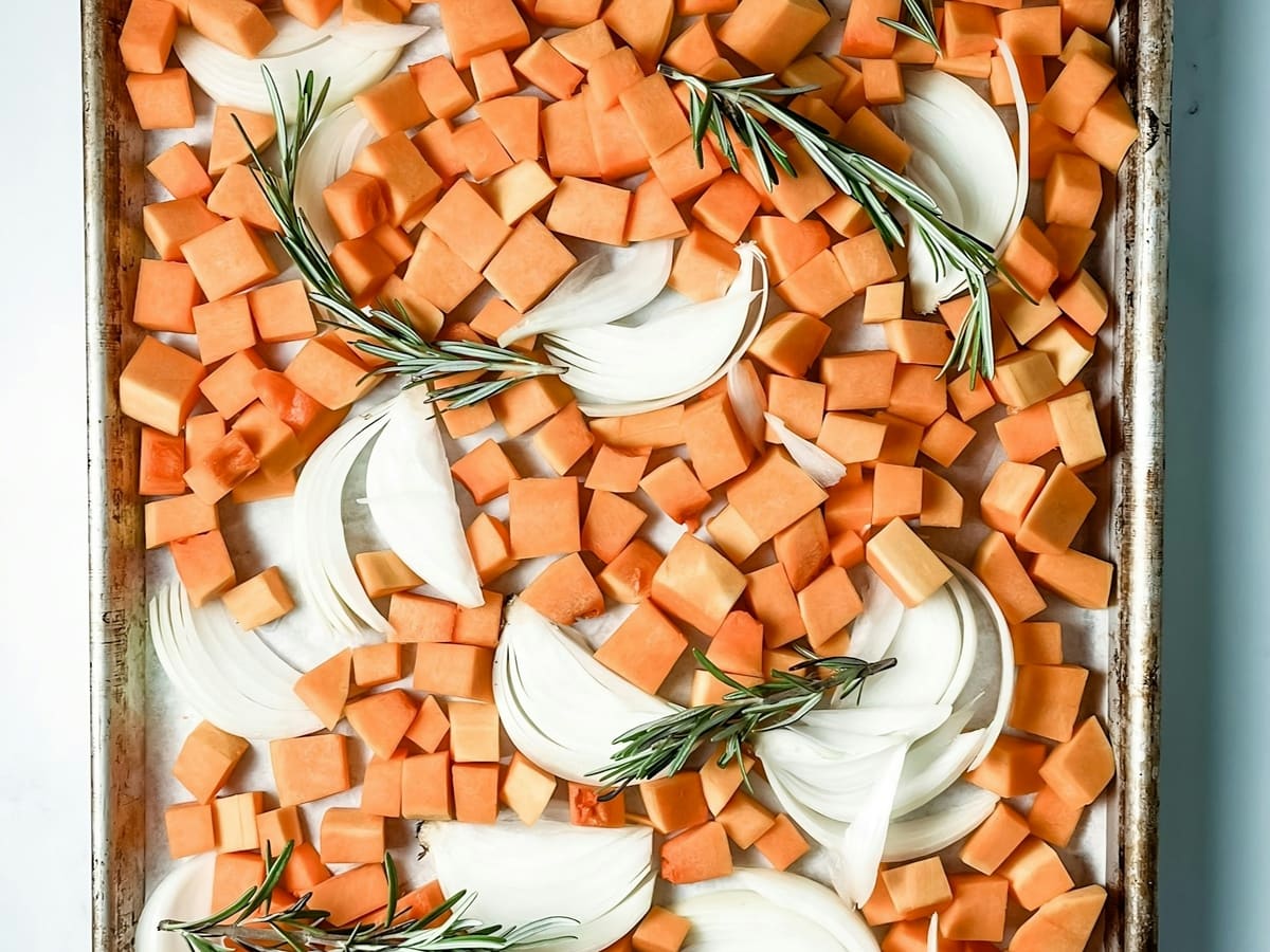 Diced butternut squash, sliced onion and rosemary sprigs spread on a parchment-lined sheet pan before roasting.