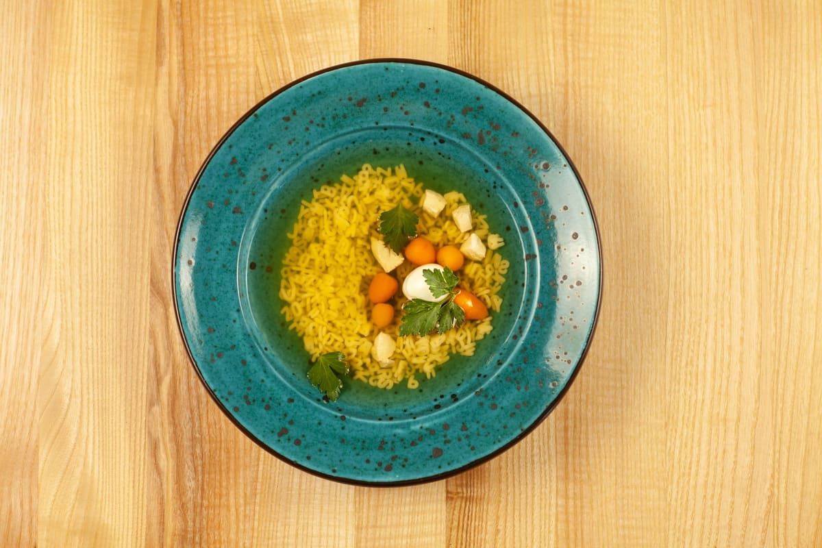 A teal bowl of clear golden broth holding tiny alphabet pasta, small pieces of carrot, and parsley leaves, on a light wooden table.