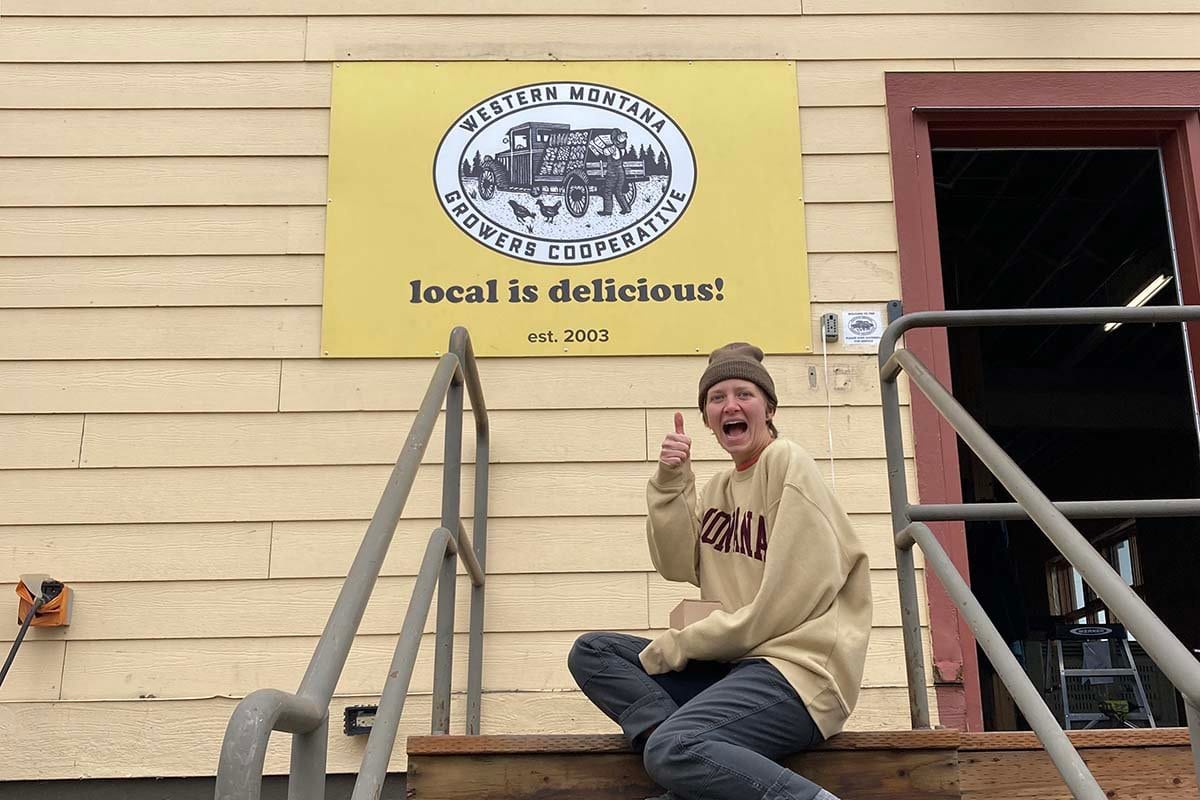A person on the steps of the Western Montana Growers Cooperative warehouse beneath its sign.