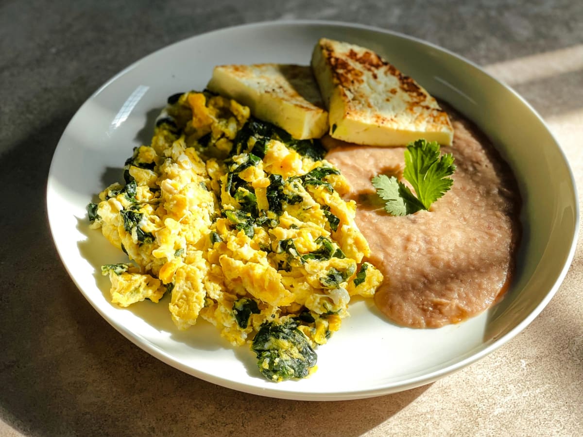 Scrambled eggs cooked with chopped spinach on a white plate, beside refried beans with a cilantro leaf and two slices of griddled white cheese.