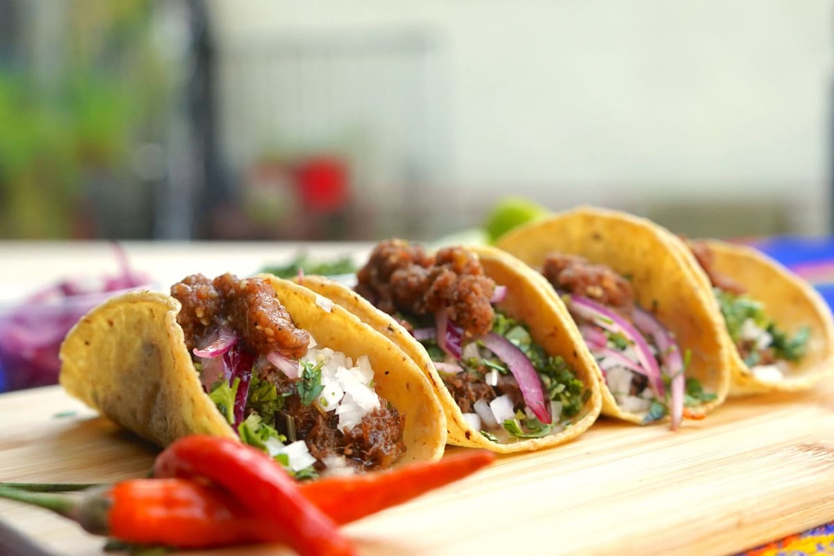 Four soft corn tortillas on a wooden board, each filled with browned crumbled meat, chopped white and red onion, and cilantro.