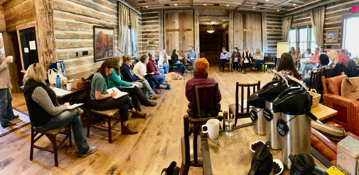 People seated in a circle, listening and sharing notes in a wood-lined meeting room.