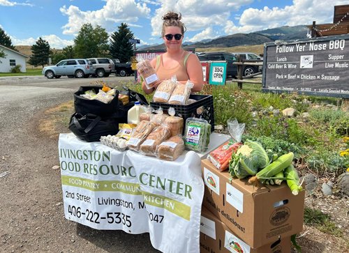 A person at an outdoor table stacked with bread, milk, eggs and vegetables under a Livingston Food Resource Center banner.