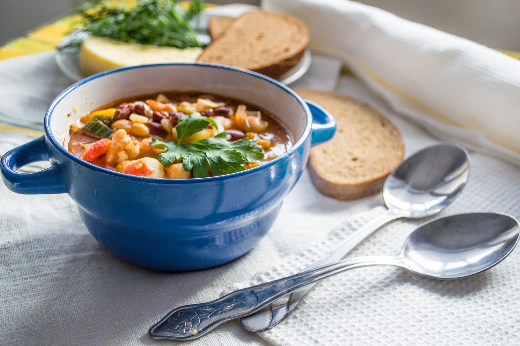 A blue pot of vegetable soup beside bread and two spoons.